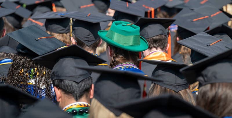 A graduate in a vibrant green fedora stands out among a crowd of other graduates wearing traditional black caps and gowns.